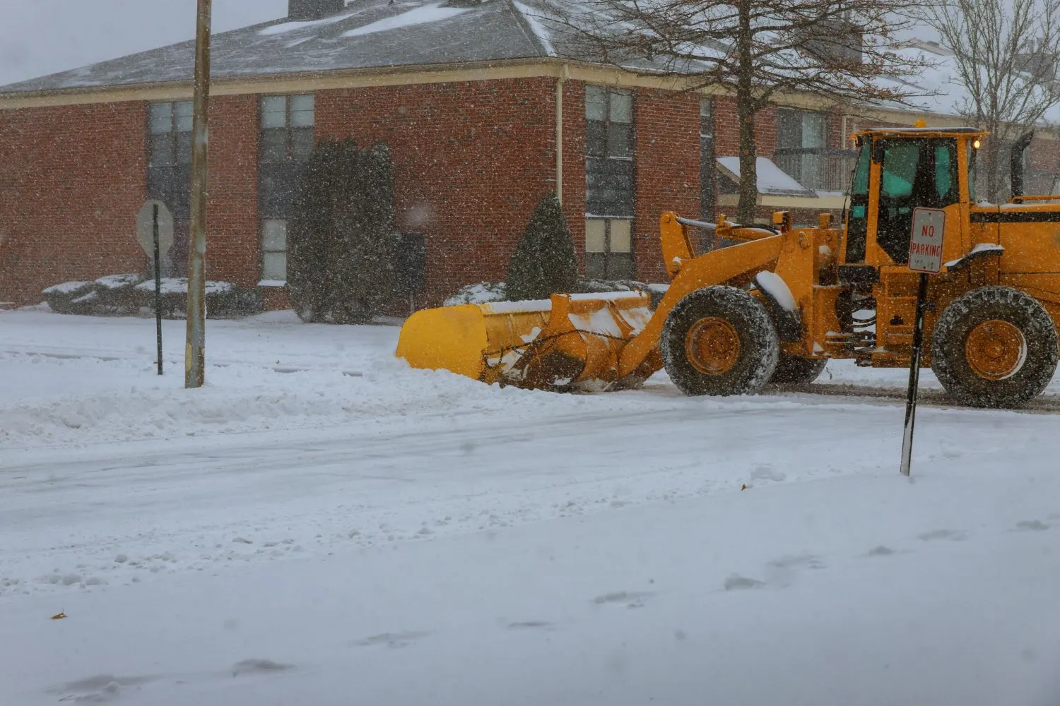 A yellow snow plow is clearing a snowy street in front of a brick building.