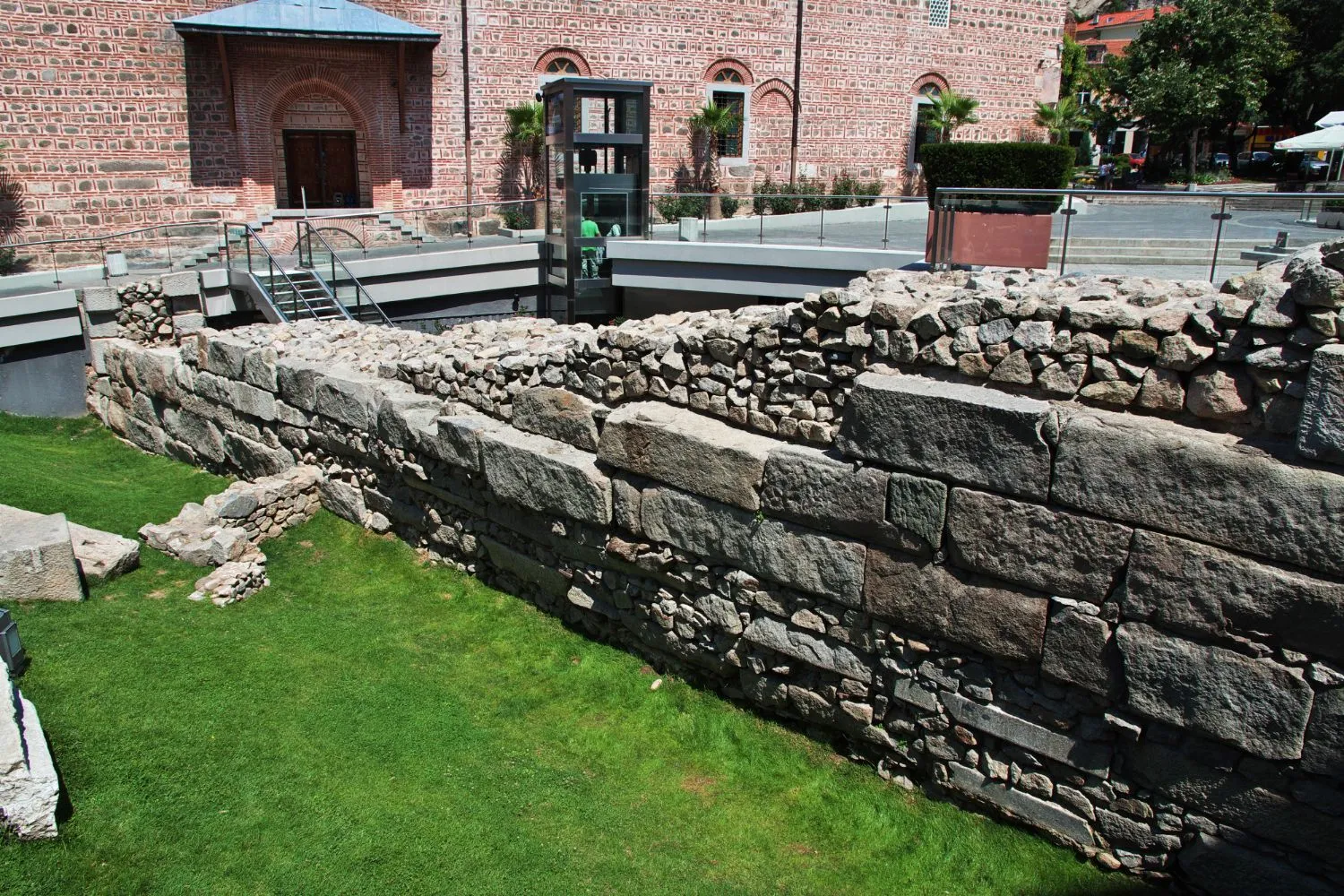 A stone wall is surrounded by green grass in front of a brick building